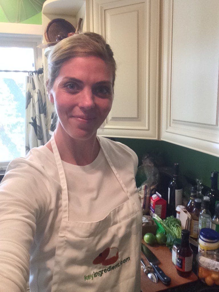 A woman in a white apron taking a selfie in a kitchen.